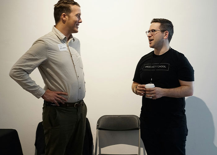 Two men talking at an event, one smiling. The other, wearing a Product School shirt, holds a coffee cup. They needed better friends.