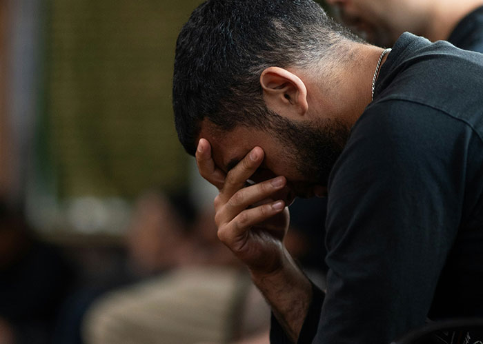 A man with dark hair and a beard, in a black shirt, covers his face with his hand, looking down in distress, symbolizing needing better friends.