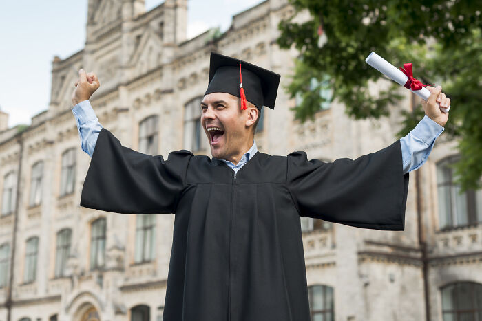 An ecstatic graduate in a cap and gown, holding a diploma, celebrates with raised arms, feeling the joy of petty revenge after years of hard work.