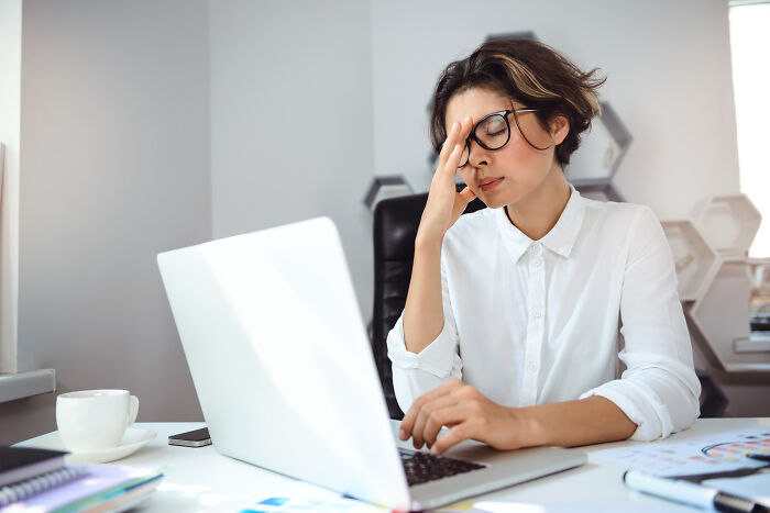 A stressed woman with glasses rubs her temples while looking at a laptop, embodying a petty revenge story.