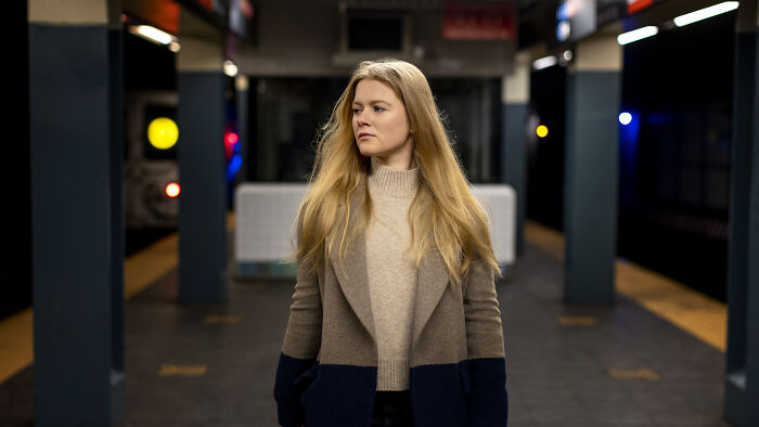A blonde woman in a stylish coat, waiting at a subway station, contemplating a petty revenge story.
