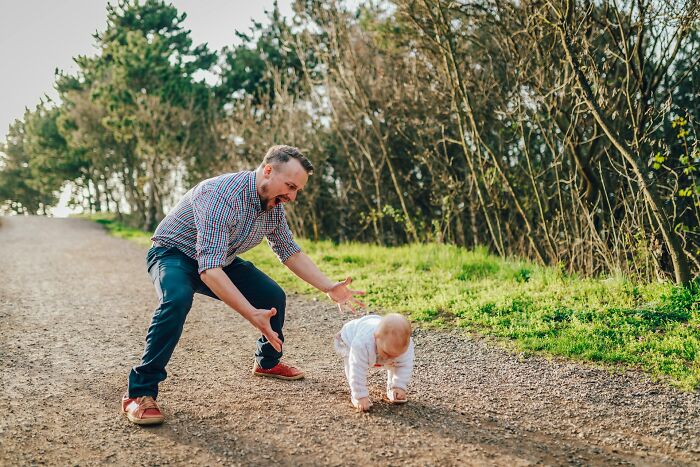 A happy dad raising daughters, encouraging his baby who is crawling on a path outdoors, surrounded by trees and grass.