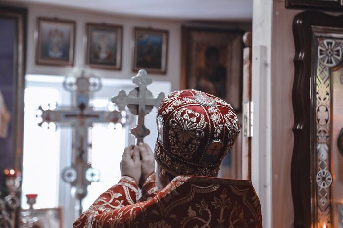A priest in ornate red and gold robes holds a cross in a church, symbolizing those who leave the Church.
