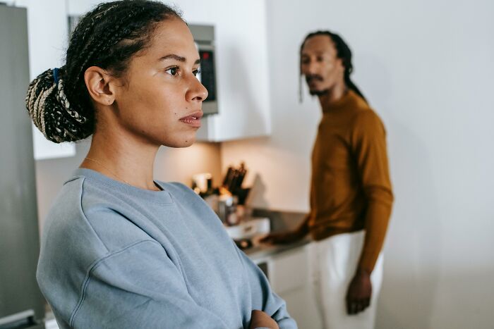 A woman with braided hair looks away, arms crossed, with a man blurred in the background, hinting at family secrets.