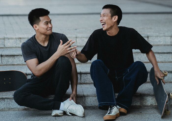 Two young men, likely brothers, sharing a laugh and discussing family secrets on concrete steps with skateboards nearby.