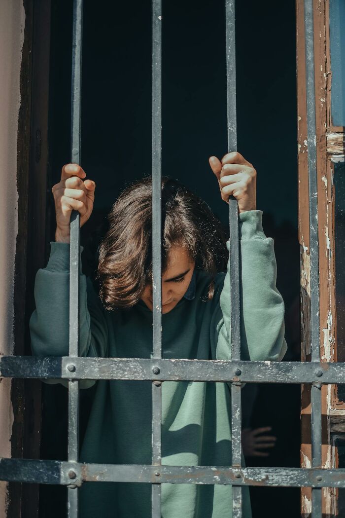 Person behind metal bars looking down, symbolizing dark family secrets and emotional confinement.