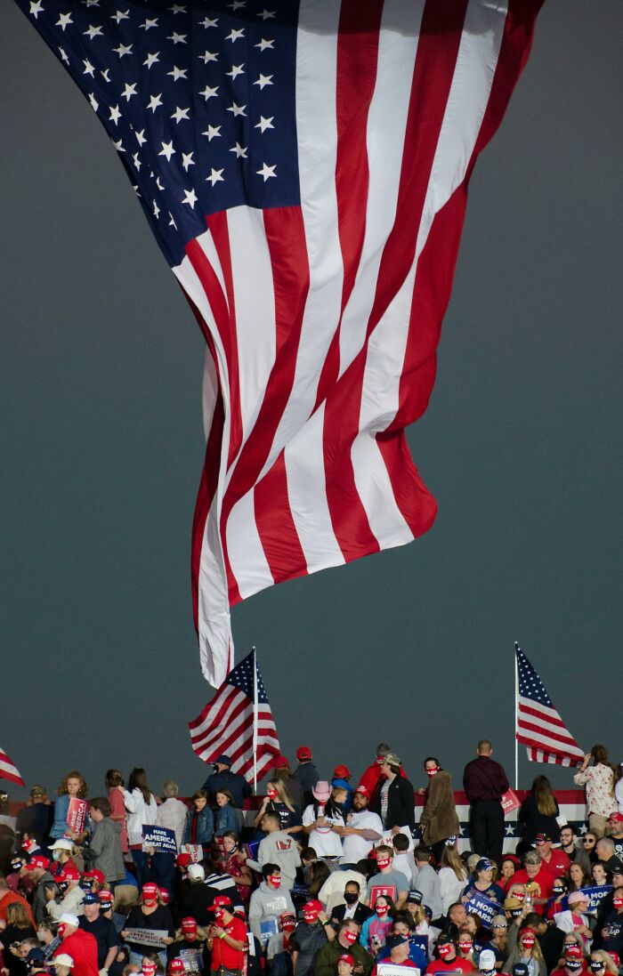 Giant American flag draped over a rally crowd against a dark sky. Attendees gather for a MAGA influencer event.