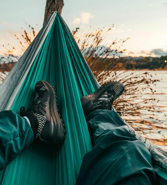 Person relaxing in a hammock by the water at sunset, wearing dark sneakers. A cheat code for surviving adult life.