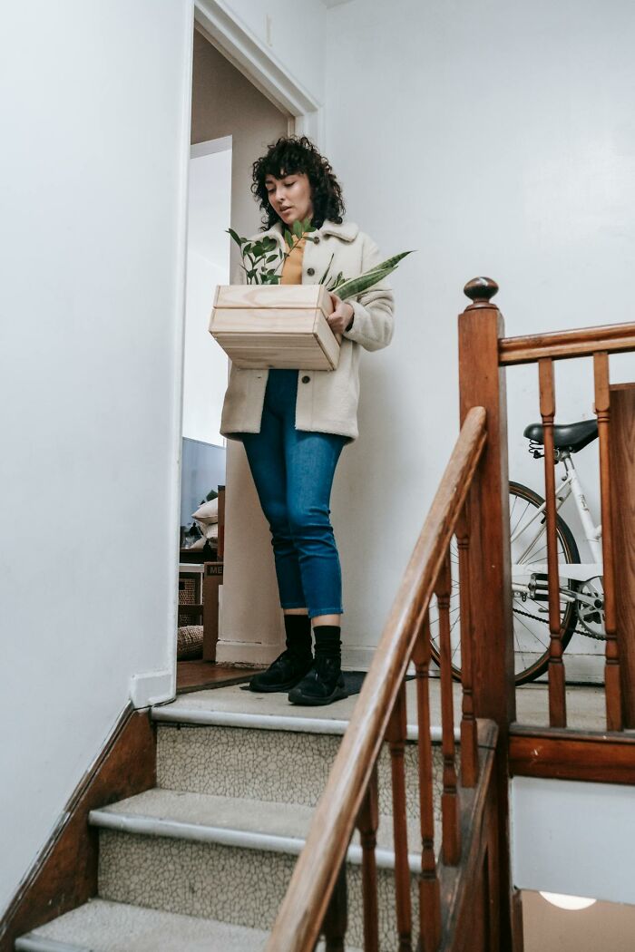 A woman in a cream jacket and jeans carries a wooden crate of plants down stairs, symbolizing moving out after divorces.