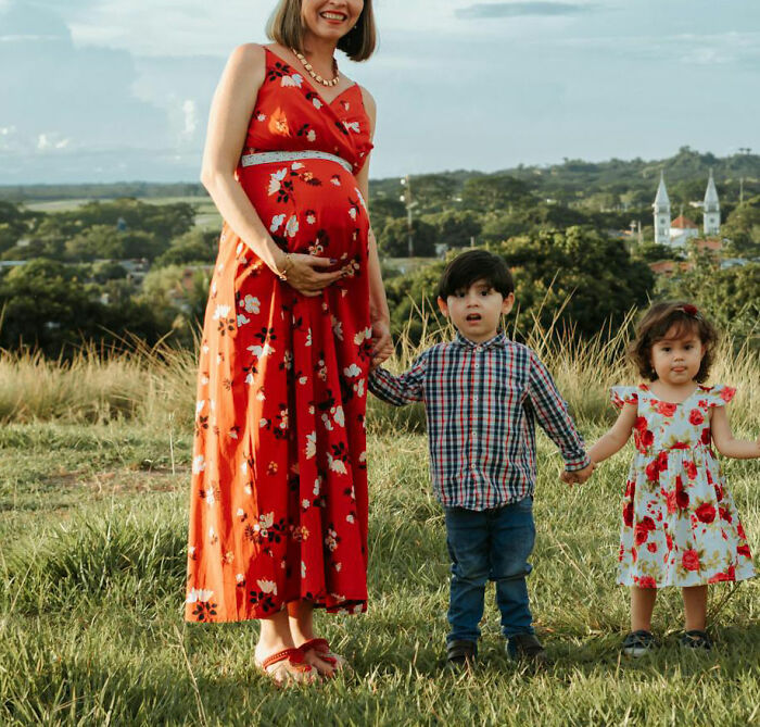 Pregnant woman in a red floral dress holding hands with two young children in a grassy field. God Bless America.