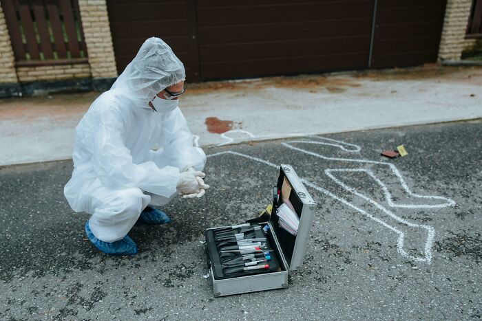 A forensic investigator in a hazmat suit examines a workplace incident scene with a chalk outline and bloodstain.