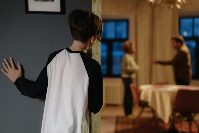 A child in a black and white shirt peers around a doorframe, observing parents arguing in the background. Family secrets.