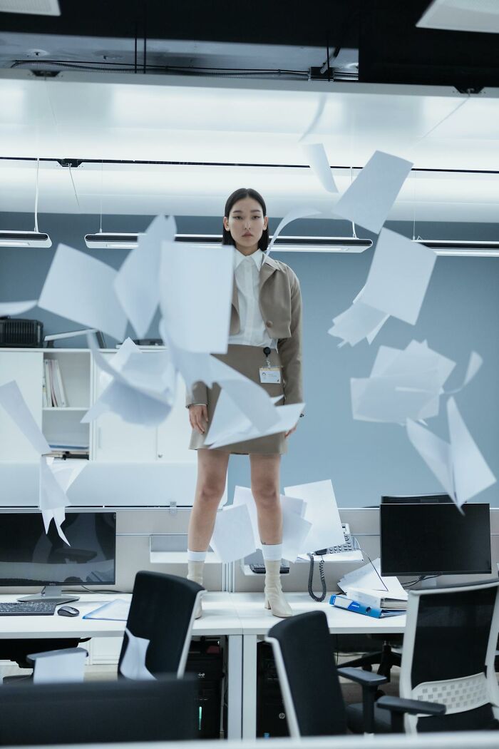 Young employee stands on desk amid flying papers in an office, capturing wild times work frustration and quitting moments.