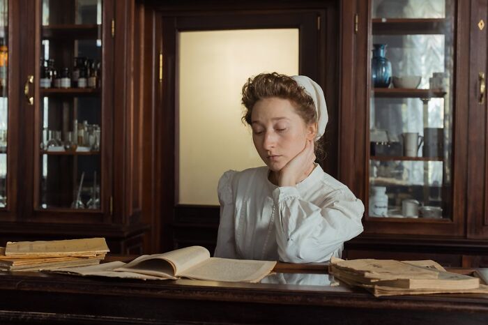 Young woman dressed as a mom sitting at a wooden table reading vintage documents in a room with glass cabinets.