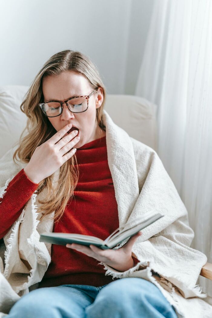 Woman wearing glasses, wrapped in a blanket, yawning while reading a book about weird medical facts.