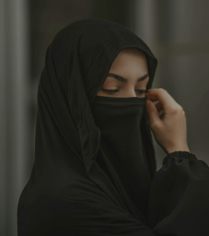 A woman in a black niqab and hijab, looking down. Her hand subtly adjusts her veil, hinting at anonymous confessions.