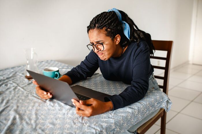 Young woman with glasses looking mortified at laptop screen after accidentally sending a message, sitting at a dining table.
