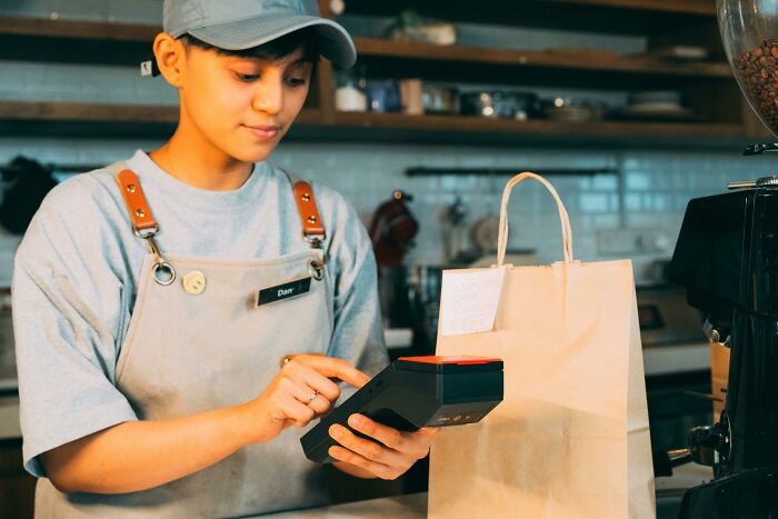 Barista in a coffee shop using a card reader machine with a takeaway bag nearby related to urban legends.