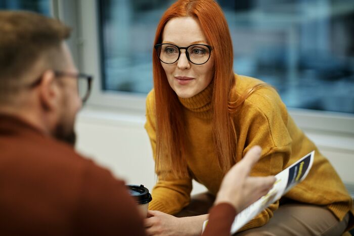 A red-haired woman with glasses listens attentively to a person holding a document, discussing cheat codes for surviving adult life.