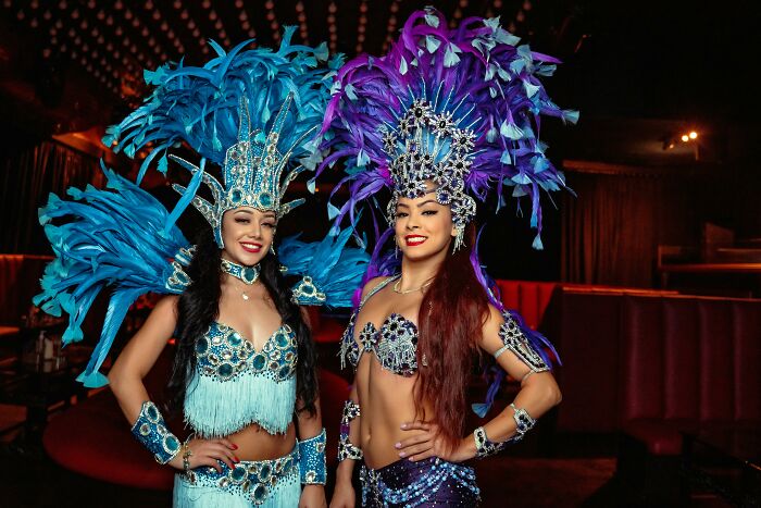 Two smiling performers in elaborate blue and purple feathered carnival costumes. Wildest rumours.