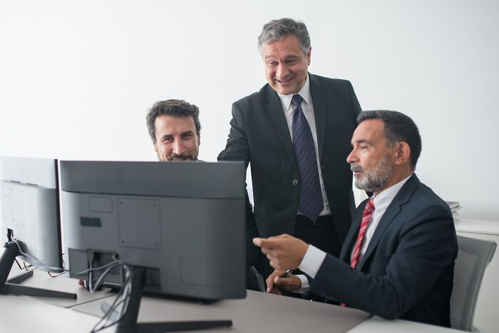 Three smiling businessmen in suits collaborate around monitors. They are discussing how they lost everything. God Bless America.