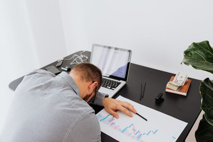 A man slumped on a desk with his head down next to a laptop, charts, and money, indicating someone who lost everything.