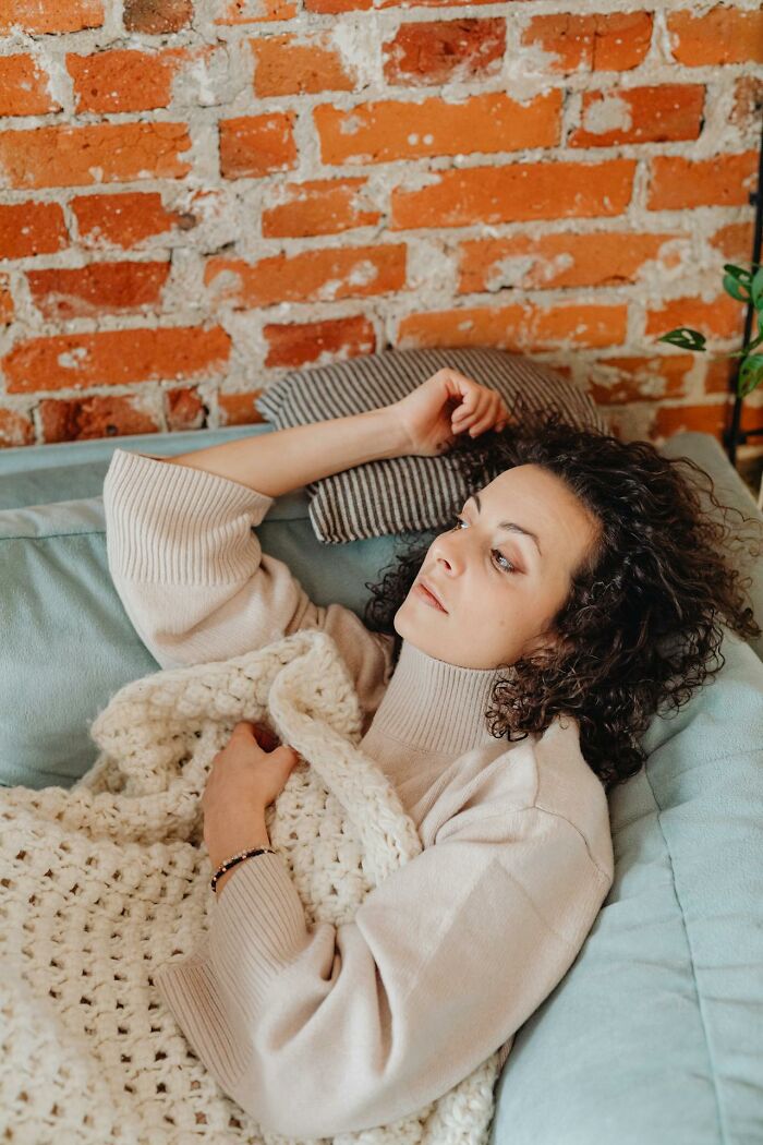 Woman lying awake on couch with blanket and pillow, appearing restless and unable to sleep, against brick wall background.