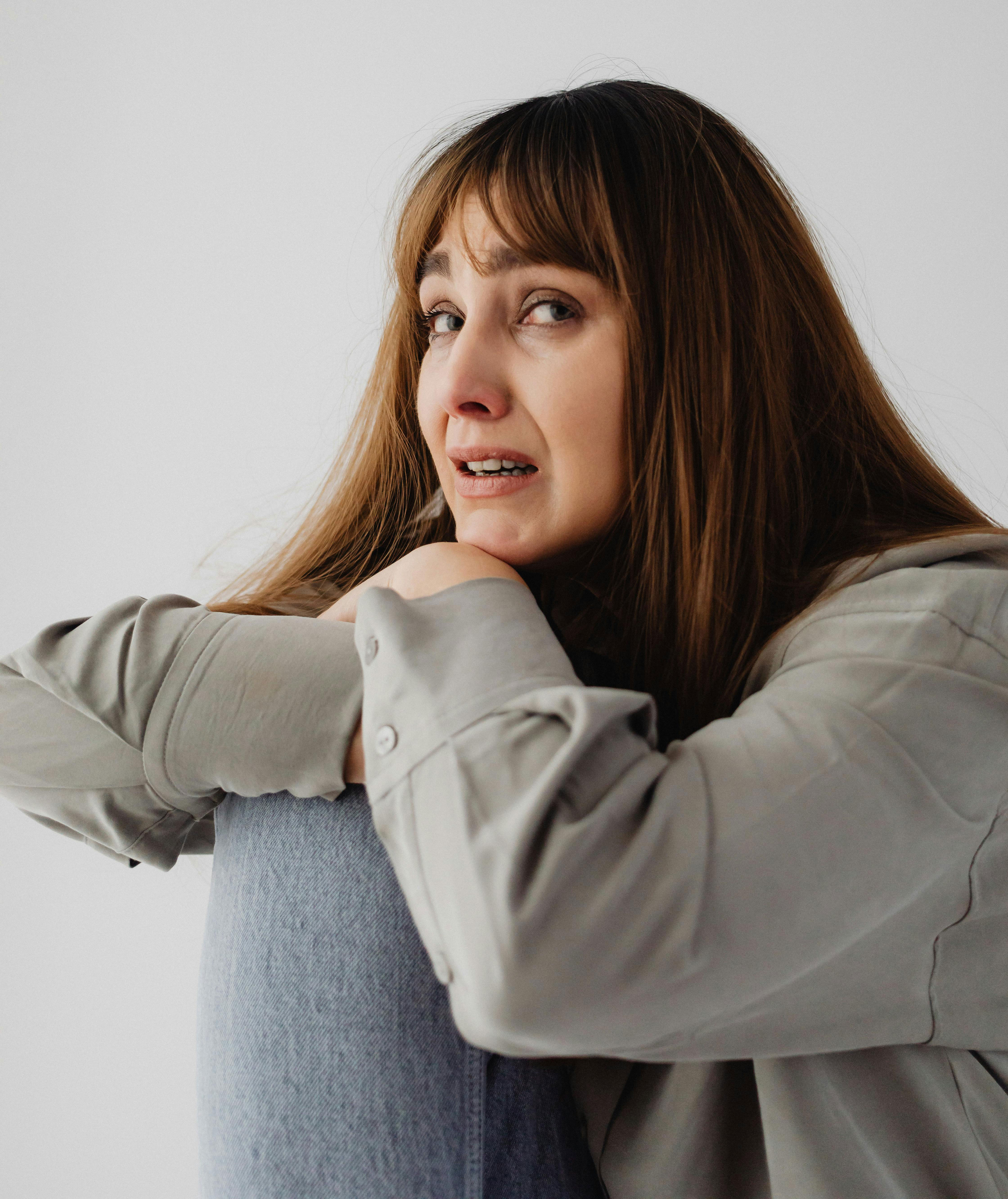 A woman with long brown hair, wearing a gray shirt and blue jeans, looking distressed and teary-eyed, facing divorce.
