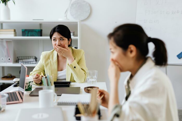 Two women, one in focus in a green blazer, both covering their mouths, showing workplace incidents and shaken reactions.