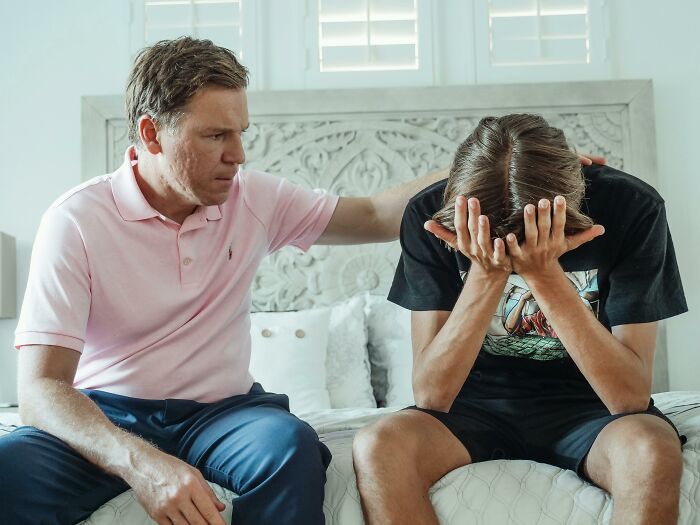 A father comforts his distressed child, who has their head in their hands, on a bed. Illustrates family secrets.