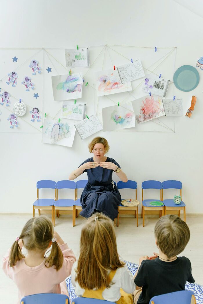 Woman teaching children in a classroom setting surrounded by colorful drawings, illustrating stories about moms.
