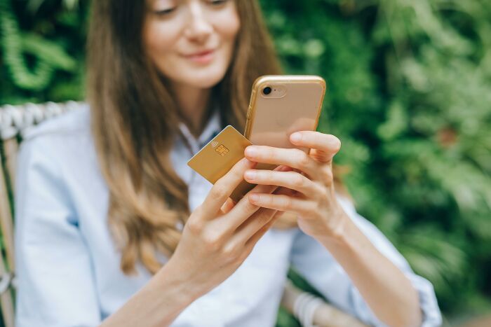 A smiling woman holding a gold credit card and a smartphone, considering a purchase or online confession.