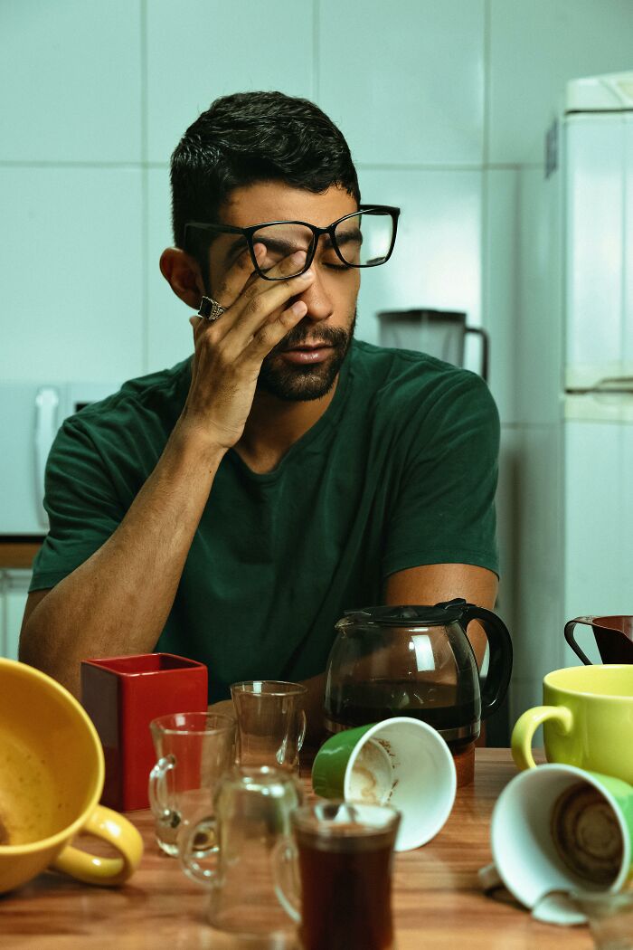 A man in glasses rubs his eye, looking tired amidst many used coffee cups. A scene portraying workplace incidents and stress.