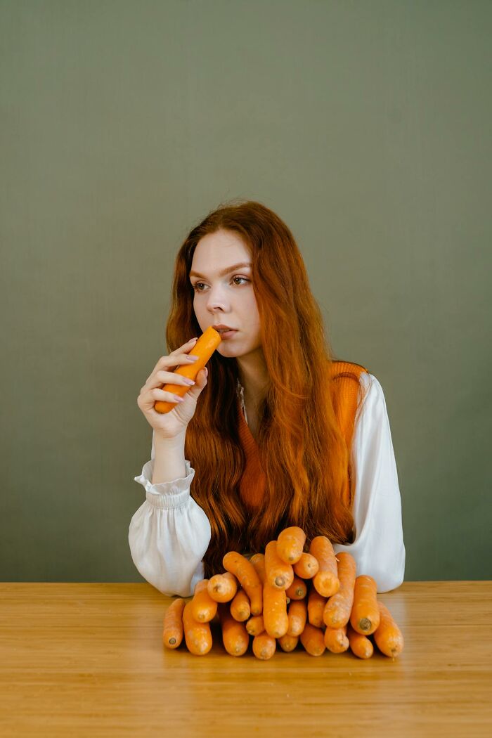 Young woman with red hair holding a carrot near her mouth with a pile of carrots on the table, medical facts concept.