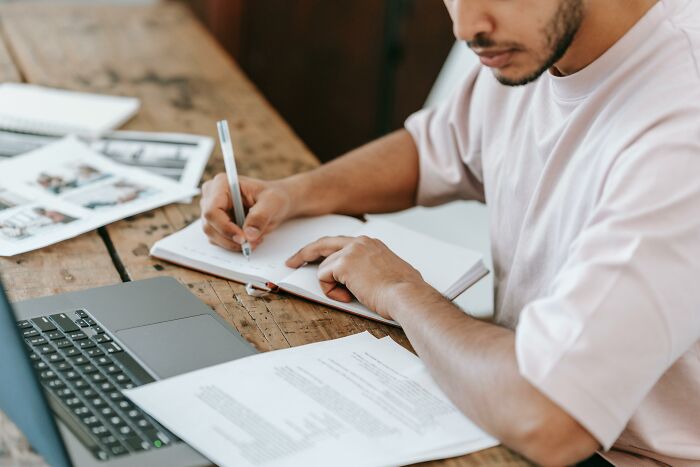 A man in a light pink shirt writing in a notebook next to a laptop. Test your general knowledge.