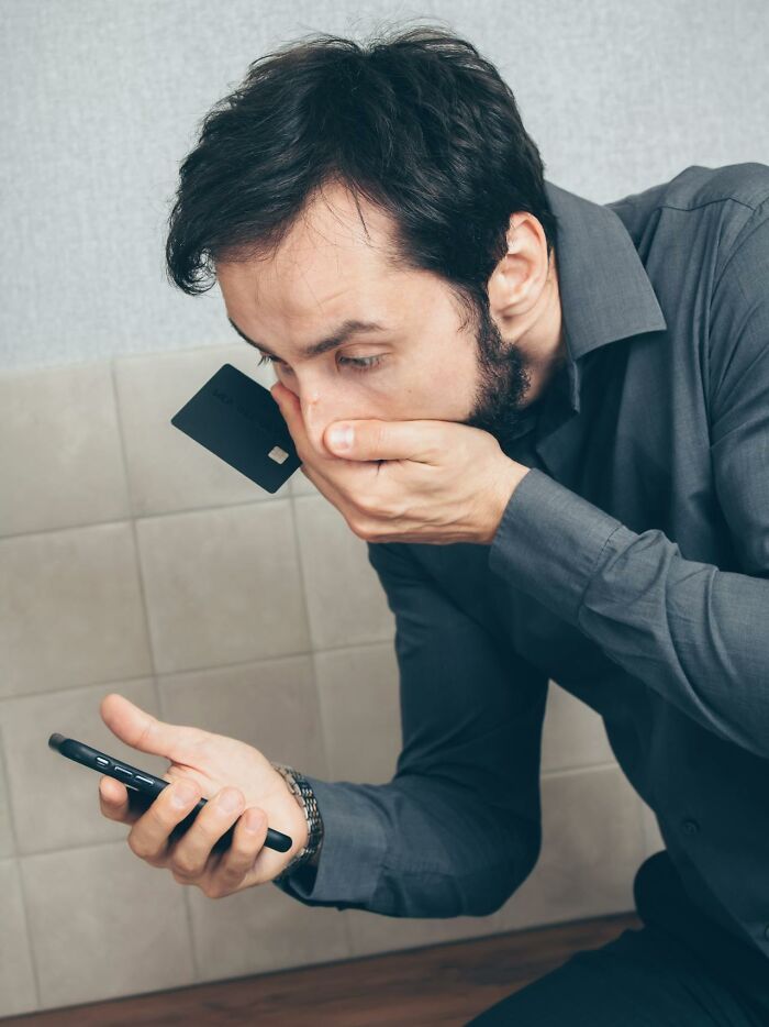 A man in a gray shirt looks distraught, covering his mouth, holding a phone and a black credit card. Represents those who lost everything.
