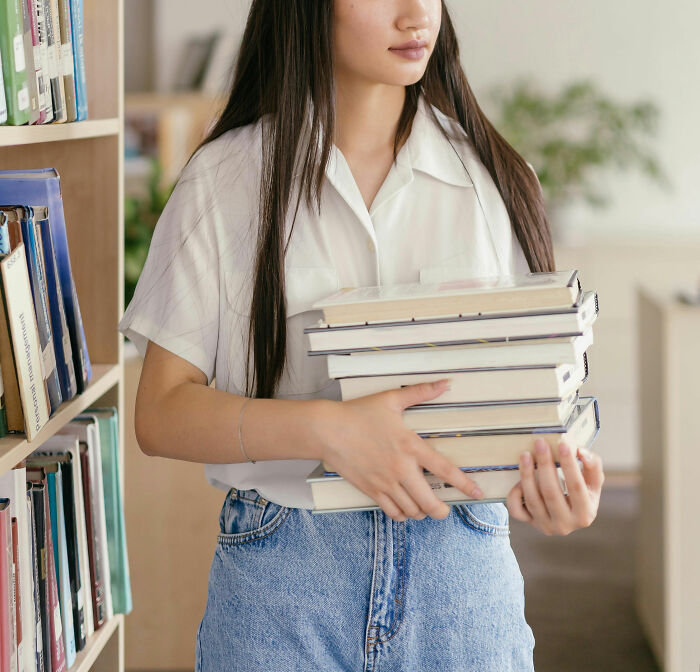 A young woman in a library, wearing a white shirt and jeans, carrying a stack of books. Exploring messy anonymous confessions.