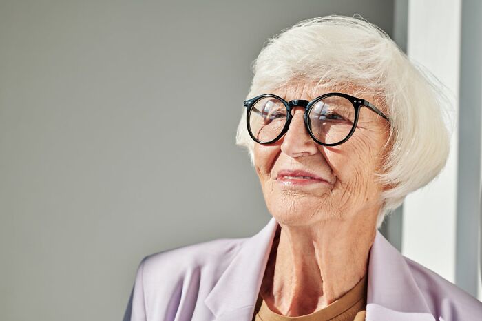 Elderly woman with white hair and glasses smiling softly, reflecting love and warmth in stories about moms.