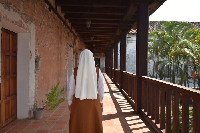 A nun from behind, walking down a sunlit corridor with wooden railings and tropical plants outside. Nuns and priests leaving the church.