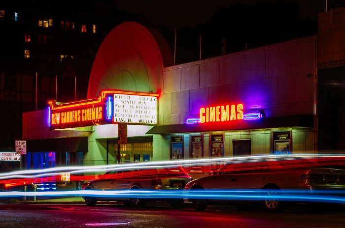 Neon-lit cinema at night with vintage car light trails, evoking a classic 60s movie scene atmosphere in an urban setting.