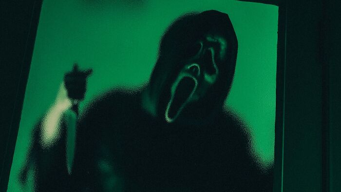 A shadowy figure in a Ghostface mask holding a knife, seen through a green textured glass, evoking horror film knowledge.