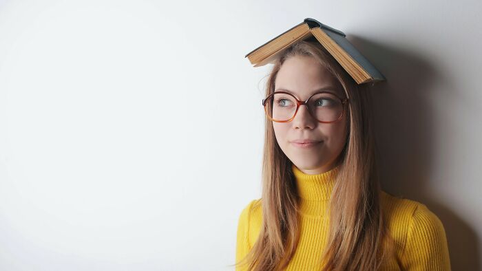 Young woman with a book on her head, wearing glasses and a yellow turtleneck, pondering advanced English words.