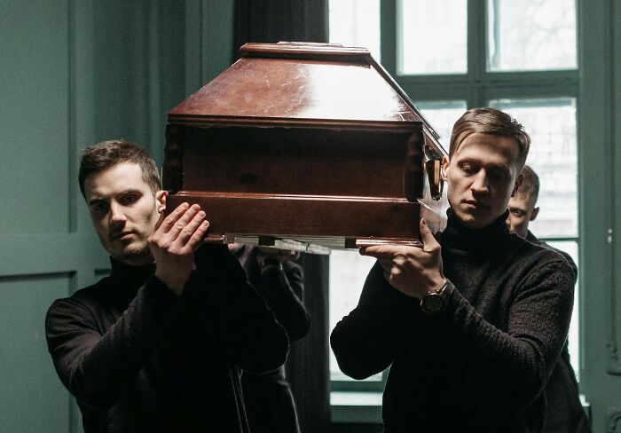 Two men in black turtleneck sweaters carrying a coffin, looking somber. This image evokes wild rumors about a secretive event.