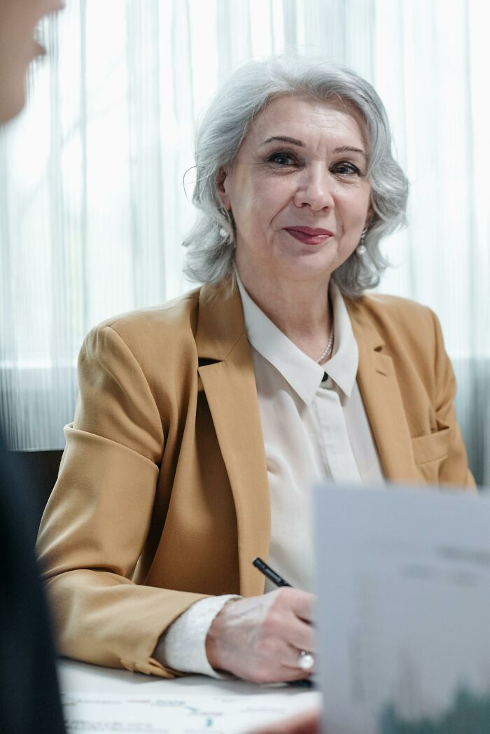 Older woman smiling gently, dressed in a tan blazer, representing inspiring and interesting stories about moms.