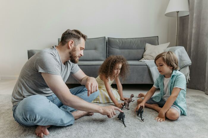 A father playing with two kids and dinosaur toys on the floor, potentially sharing family secrets.