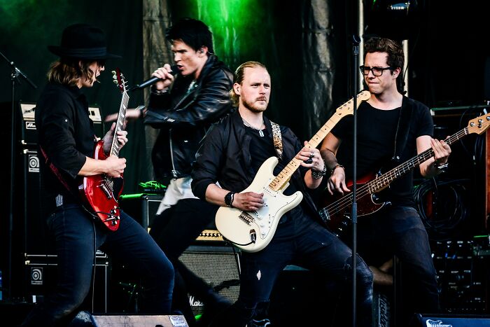 A rock band performs live, guitars and bass, under green lights. Secrets are part of the core performance.
