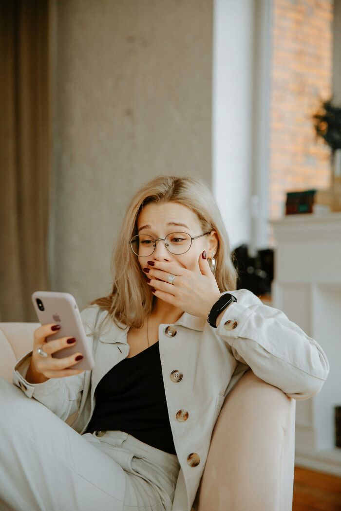 Blonde woman in glasses looking shocked at her phone, hand over mouth, revealing family secrets.