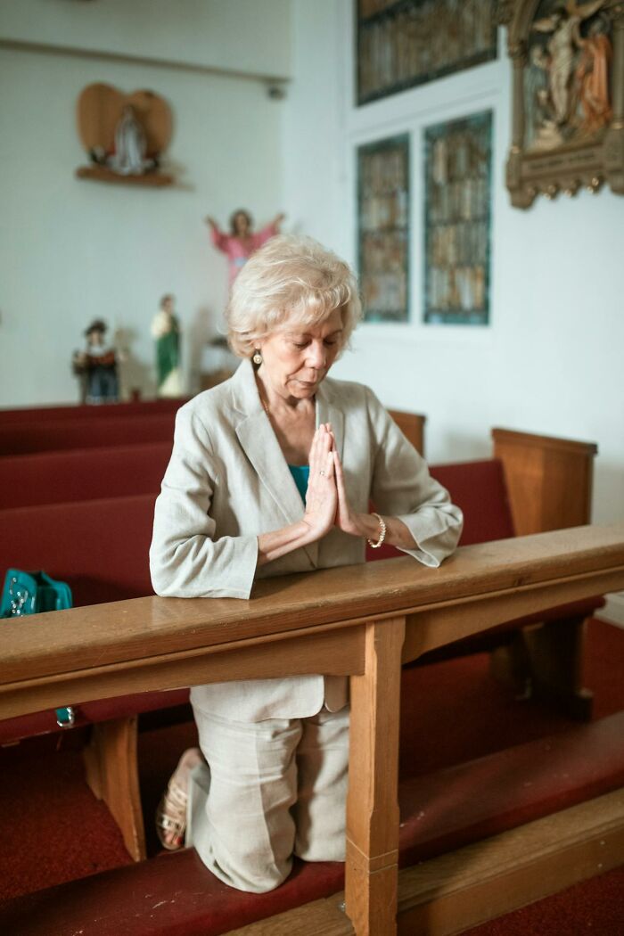 An older woman with blonde hair, dressed in a pantsuit, kneels and prays in a church, reflecting on losing everything.