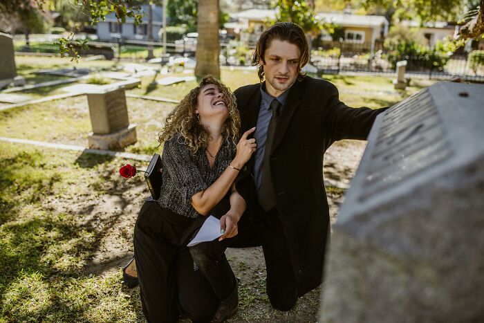 A distraught woman with curly hair and a man in a black suit kneeling by a tombstone. Disturbing sounds of grief.