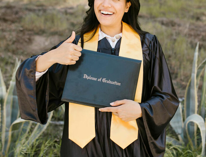 Happy graduate woman holding diploma and giving thumbs up outside, celebrating achievements and stories about moms.
